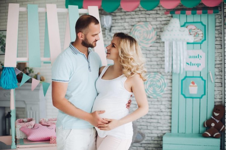 Sesja ciążowa – przyszli rodzice w pastelowej scenerii, mężczyzna obejmuje brzuszek kobiety