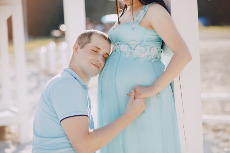 Mężczyzna przytula głowę do brzucha ciężarnej partnerki w błękitnej sukni na plaży