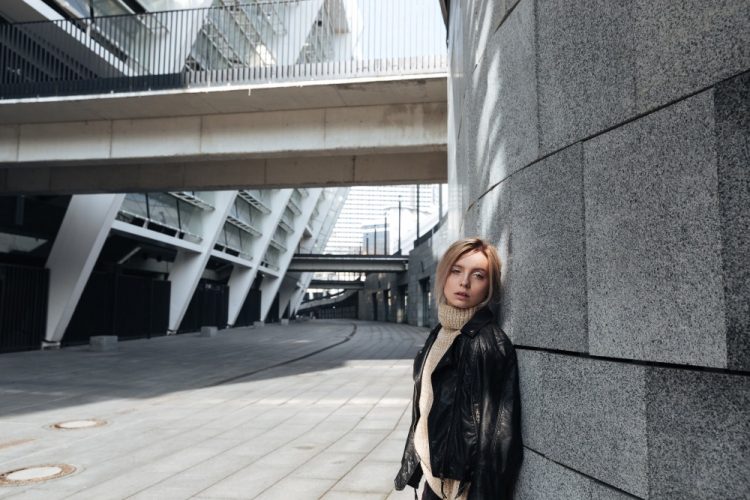 Kobieta w skórzanej kurtce i golfie pozuje przy nowoczesnym budynku z betonu i szkła