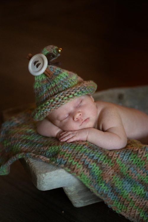 Noworodek śpiący w drewnianej misie, ubrany w zielono-brązową czapeczkę z ozdobami – sesja niemowlęca