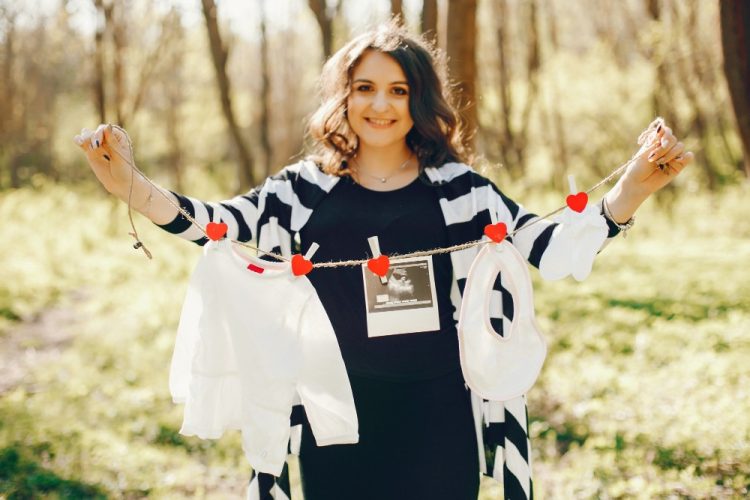 Uśmiechnięta kobieta w ciąży trzyma sznurek z ubrankami niemowlęcymi i zdjęciem USG podczas sesji w plenerze