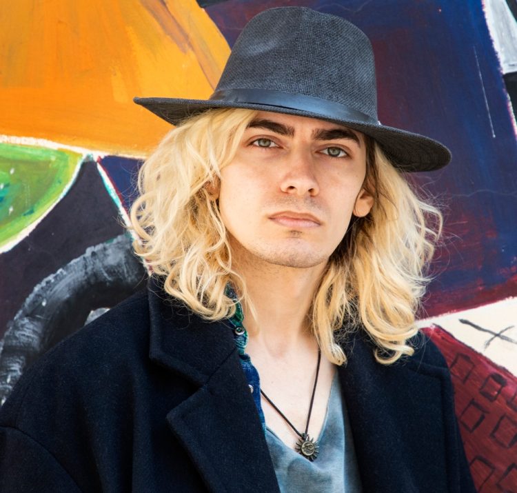 Mężczyzna z długimi blond włosami w kapeluszu i czarnym płaszczu na tle kolorowego graffiti