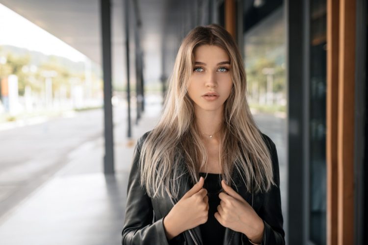 Młoda kobieta z długimi blond włosami w czarnej skórzanej kurtce, pozująca na tle miejskiej architektury