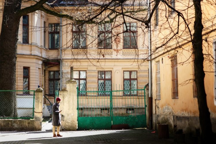 Mężczyzna w płaszczu i czerwonej czapce spacerujący po podwórku kamienicy w słoneczny dzień