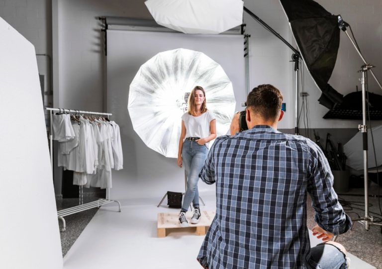 Fotograf robiący zdjęcie modelce w białej koszulce i dżinsach, w profesjonalnym studio fotograficznym z blendą i lampam